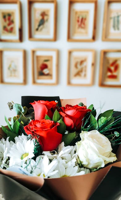 A close-up of a floral arrangement featuring vibrant red roses and white flowers with lush green leaves, wrapped in beige and black paper. The bouquet is held at an angle, showcasing the fresh, textur