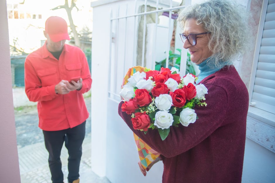 A woman with curly gray hair and glasses, dressed in a maroon sweater over a blue turtleneck, is holding a bouquet of fresh red and white roses, wrapped in colorful patterned paper. She is standing ne