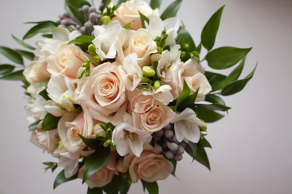 A close-up of a floral arrangement featuring pale blush pink roses, white freesias, and small green buds, all surrounded by lush green leaves. The bouquet is neatly arranged with the roses and freesia