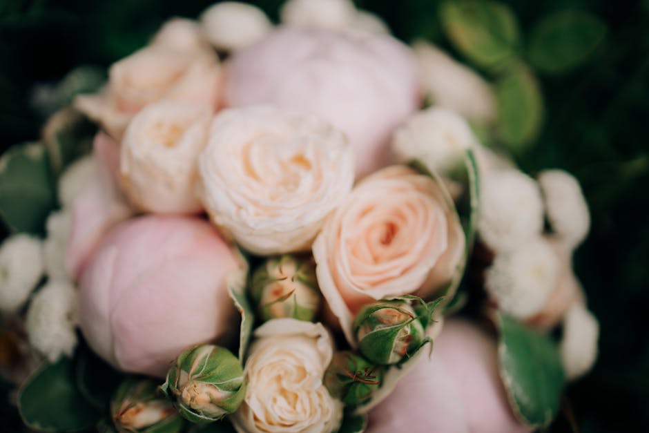 A close-up view of a floral arrangement featuring soft pink and cream roses with gently ruffled petals, complemented by small white blossoms and lush green foliage. The bouquet is arranged with a natu