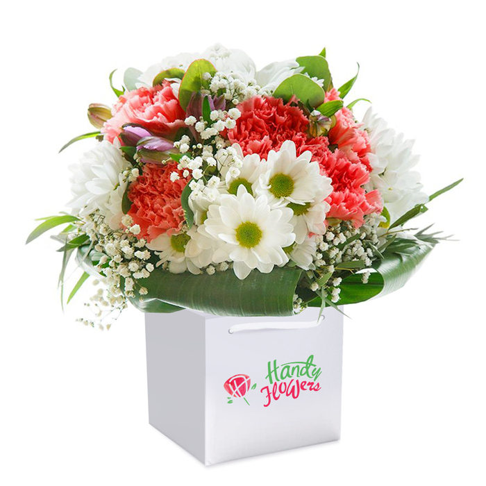 This exuberant bouquet, expertly arranged by a Kenton florist, stands upright within a radiant white square gift bag, beautifully showcased against a luminous clean background. Accents of coral and peach carnations, whose softly ruffled petals ripple with rich texture, harmonise brilliantly with bursts of pristine white daisies, their striking chartreuse centers inviting a sense of optimism reminiscent of the area's springtime blooms along Kenton Court. Wispy clusters of white baby's breath float elegantly throughout, softening the entire arrangement and infusing it with a gentle, cloud-like airiness. Nodding on the left, unopened buds reveal shades of purple and pink, suggesting the promise of future blossoms. Lustrous deep green foliage and wide, glossy leaves envelop the bouquet's base, grounding the structure with a polished, natural border reminiscent of Kenton's leafy garden squares. The sturdy white handles of the gift bag ensure a seamless gifting experience, whether strolling down Kenton Road or making a heartfelt delivery across Harrow. Every bloom has been placed with the attentive artistry of a florist deeply familiar with the local community, making this bouquet an elegant choice for birthdays, anniversaries, or cherished "just because" moments. The fresh presentation and balanced colours ensure this arrangement forms a memorable centrepiece for any Kenton home.