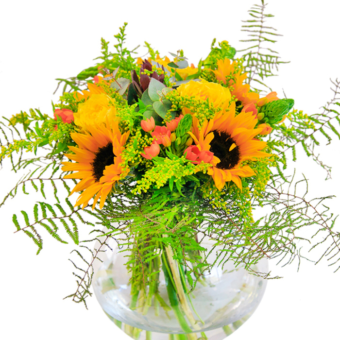 This sunlit bouquet, expertly designed by Kenton's florists, embodies a beautiful blend of warmth and joyful energy. Bright sunflowers with velvety brown centres burst into view, their golden petals arching outward like rays stretching across St. Leonard's Avenue. Nestled around these luminaries, yellow garden roses bloom in generous spirals, carrying a sense of classic charm. Glossy, deep red Hypericum berries gleam among the bouquet's foliage, their playful spark providing eye-catching contrast to the soft yellows. A lush tapestry of greenery-arched fern fronds, wispy solidago with petite yellow buds, and slender spikes-surrounds the main blooms, mimicking the layered beauty of Kenton's own gardens. Subtle bluish eucalyptus leaves provide a breezy accent, their unique tint enhancing the rustic yet refined feel. The clear glass, fishbowl-shaped vase cradles each stem, presenting the bouquet as a work of living art and inviting viewers to admire nature's detail up close. A gentle garden scent rises from the arrangement: fresh and lightly perfumed, reminiscent of a Kenton morning in bloom. This is an ideal centrepiece for celebrations, anniversaries, or simply to express happiness-every detail crafted with care and local pride.