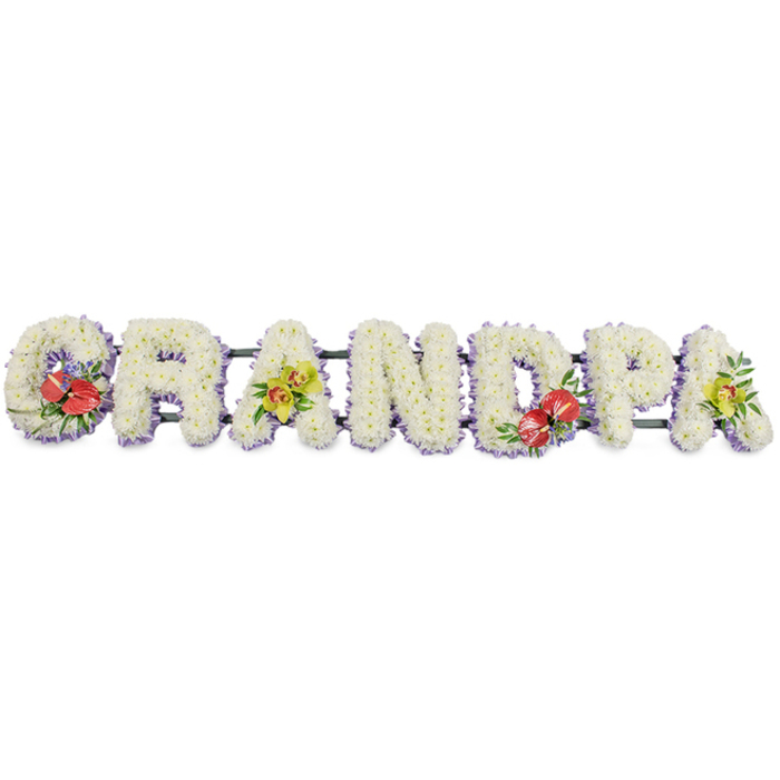 In this image, a bespoke memorial flower arrangement spells out "GRANDPA" in striking block capital letters, laid out in a perfectly straight horizontal line as if ready to be placed by the hearse or inside a Kenton funeral chapel. The entire tribute is set against a bright white background, allowing the shapes and textures to stand out with clarity. Each letter is fully covered with small, uniform white chrysanthemums, their tightly packed blooms creating a dense, cushion-like effect that feels both soft and substantial, symbolising purity and calm reflection. Along the base of every letter runs a neat band of pale lavender ribbon, providing a delicate contrast that gently defines the lower contours and adds a refined, finished look. Dark green structural bars run along the back of the word, largely concealed, ensuring the letters sit upright and aligned. To punctuate the serene white surface, four letters include thoughtfully placed sprays of brighter flowers. The 'G' on the left is embellished with glossy deep red anthuriums, their heart-shaped, waxy petals and yellow spadices surrounded by rich green foliage, drawing attention and offering a visual expression of deep love. The first 'A' features a cluster of yellow-green orchids, their slender petals and buds arranged among leafy greens to give an airy, elegant touch. Slightly right of centre, the 'D' repeats the red anthurium motif, balancing the design. On the far right, the last 'A' mirrors the first with further yellow-green orchid blooms and fresh greenery. This GRANDPA tribute, similar to the personalised designs created by Flowers Kenton for services in local churches and crematoria, provides a heartfelt, visually striking way to honour and remember a cherished grandfather.